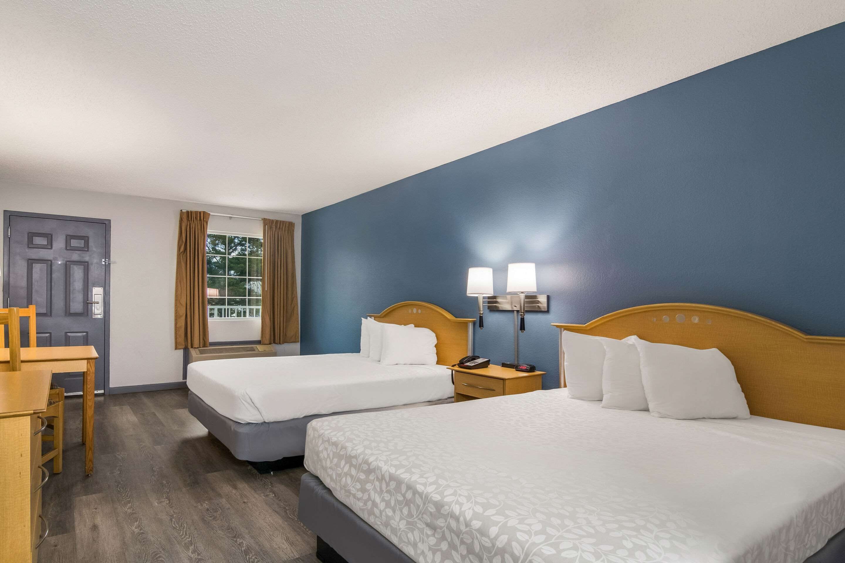A hotel room with two beds, a desk, a chair, a window with curtains, and a door.