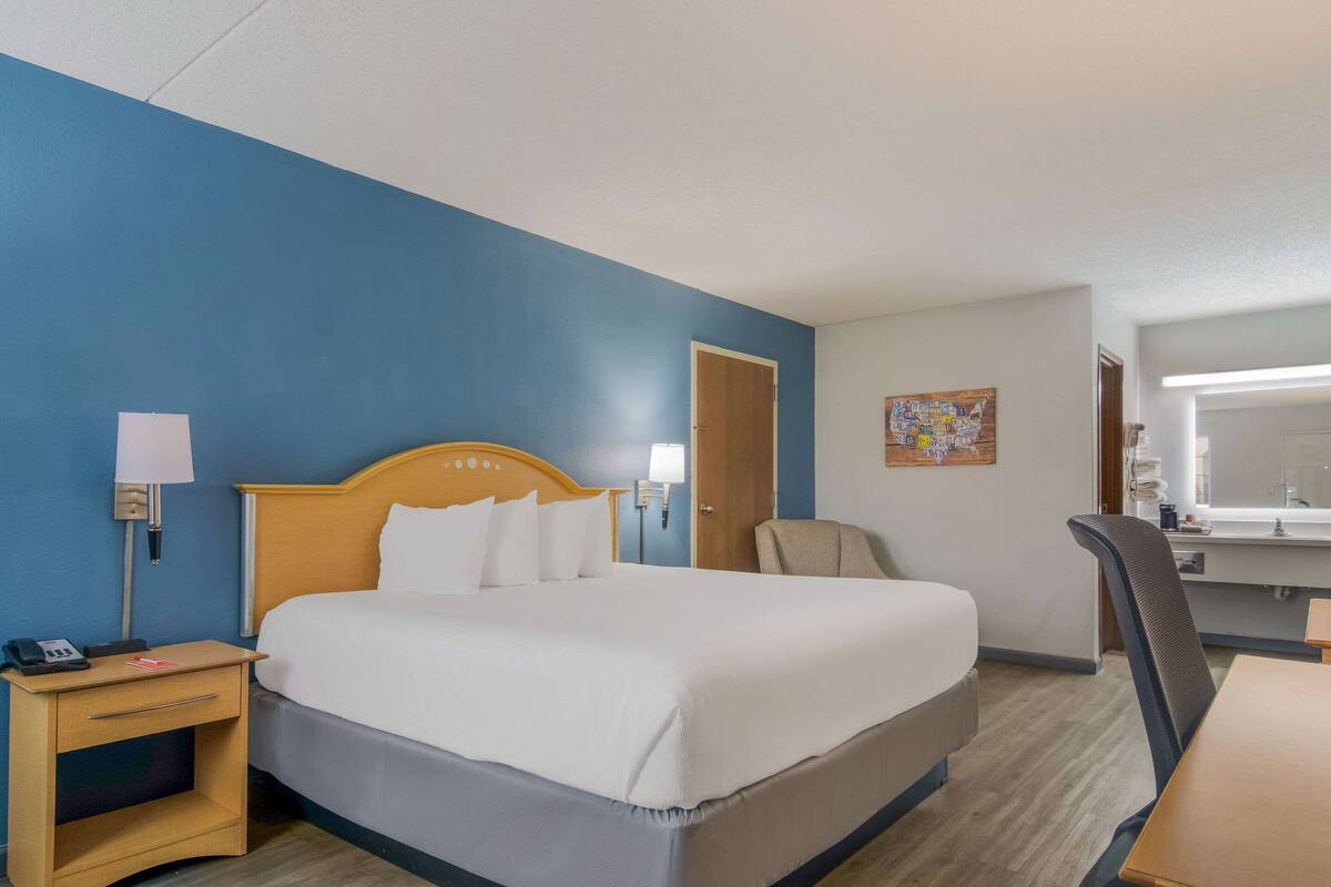 A hotel room with a bed, bedside table, lamp, chair, desk, and a bathroom mirror.