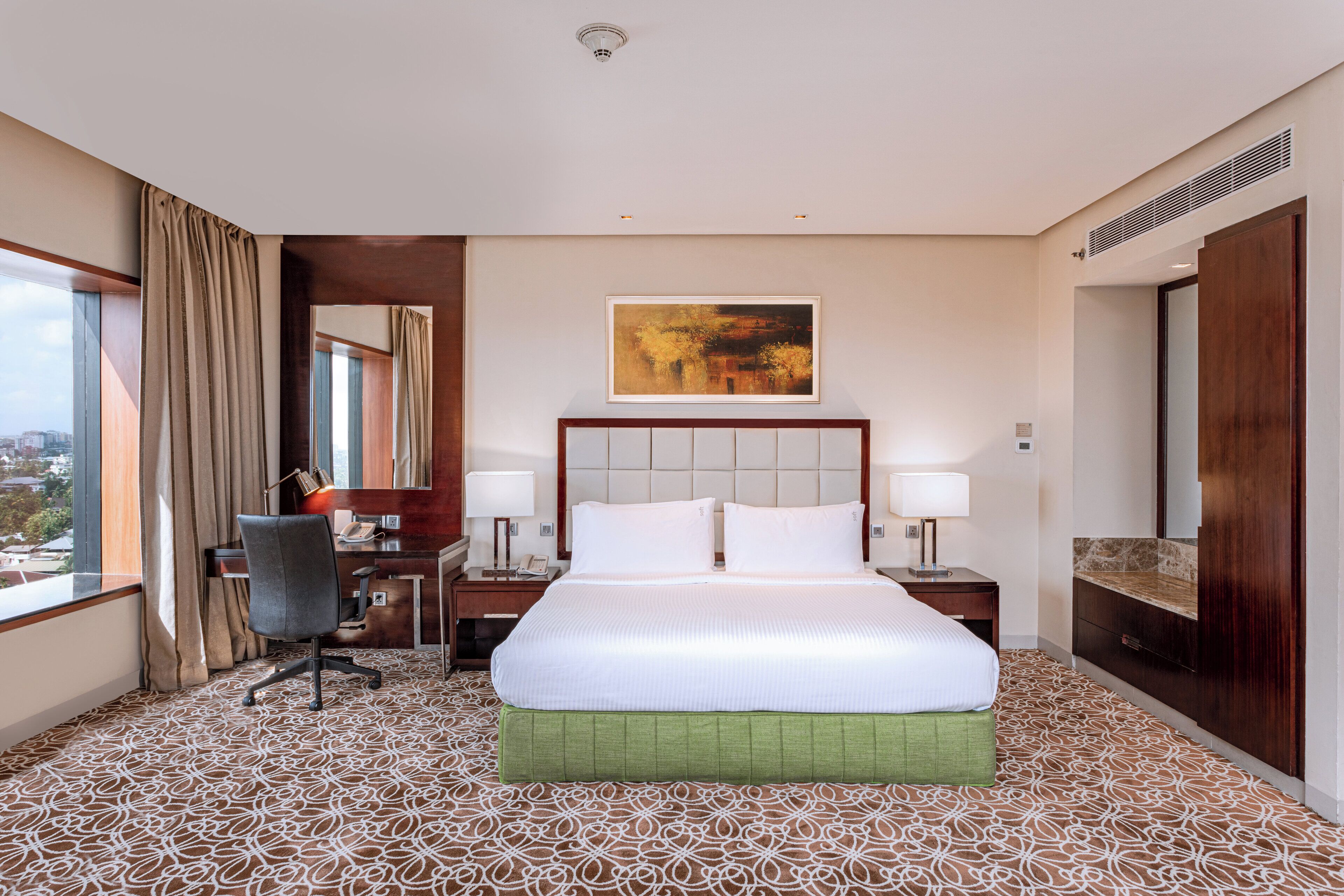 A hotel room with a large bed, a desk, and a chair. There is a window with curtains, a painting on the wall, and a bathroom with a mirror and sink.