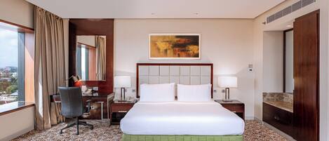 A hotel room with a large bed, a desk, and a chair. There is a window with curtains, a painting on the wall, and a bathroom with a mirror and sink.