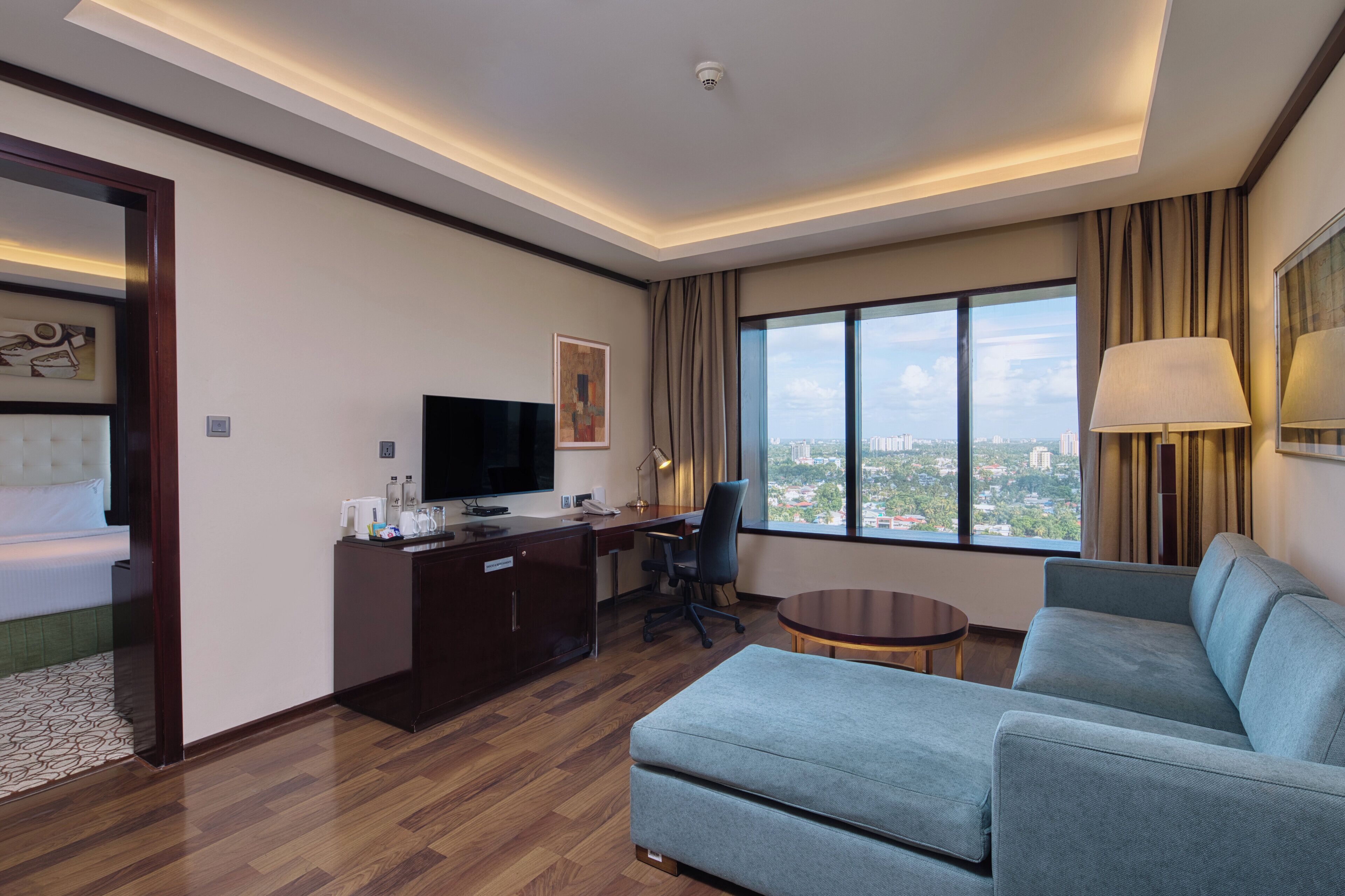 A modern hotel room with a large window, a sofa, a desk, and a television.