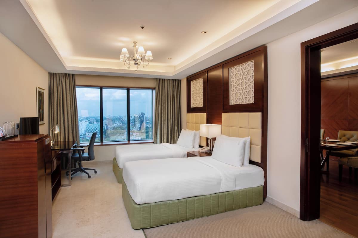 Ein Hotelzimmer mit zwei Betten, einem Schreibtisch mit Stuhl, einer Schreibtischlampe, einem Telefon und Blick auf die Stadt durch große Fenster.