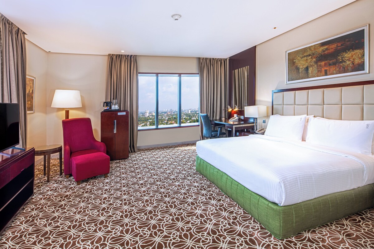 A hotel room with a large bed, a desk with a chair, a television, and a view of the city through large windows.