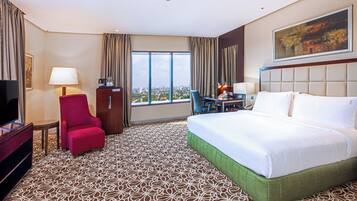 Habitación de hotel con una cama grande, un escritorio con silla, televisión y vistas de la ciudad a través de amplias ventanas.