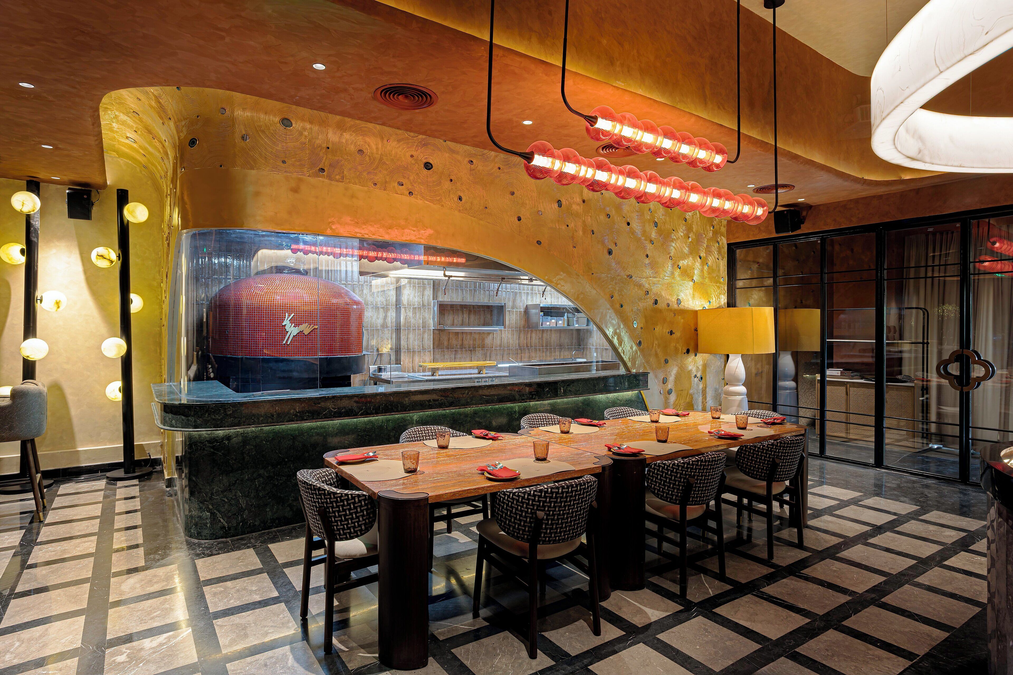 Een restaurant met een opvallend gebogen plafond, een lange eettafel met stoelen en uitzicht op de keuken door een glazen scheidingswand.