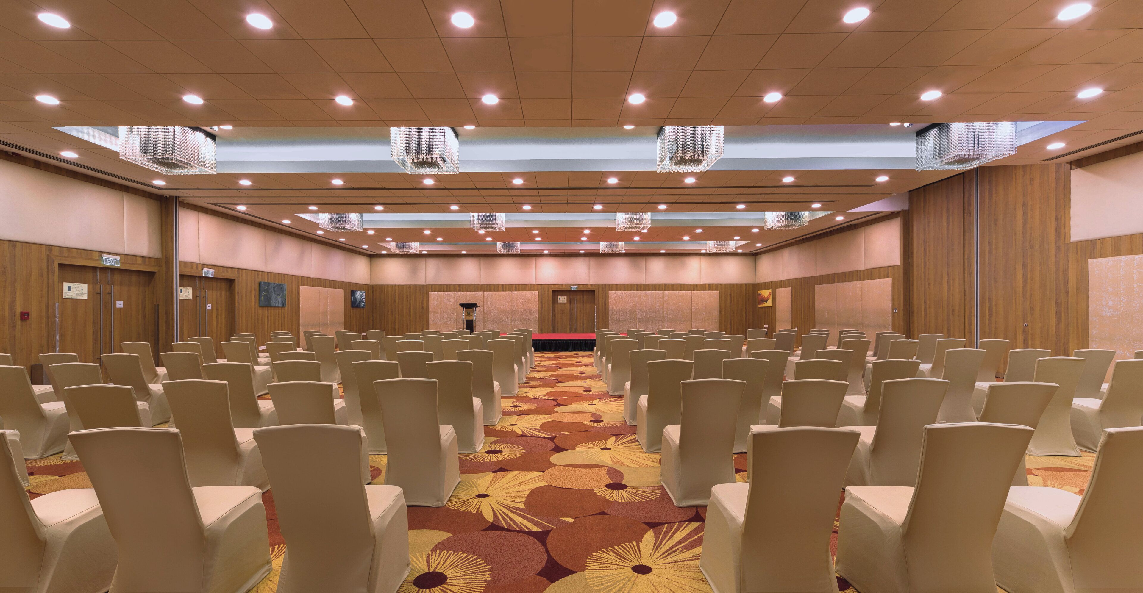 Un salón de conferencias con filas de sillas, alfombra estampada, paneles de madera y luces en el techo.