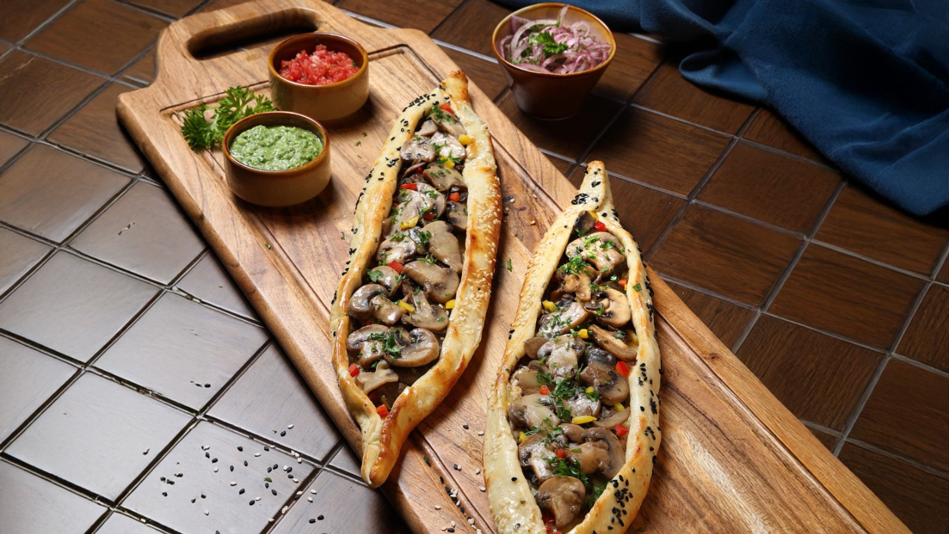 Two stuffed bread rolls on a wooden board with sesame seeds, accompanied by two small bowls of sauces and pickled vegetables.