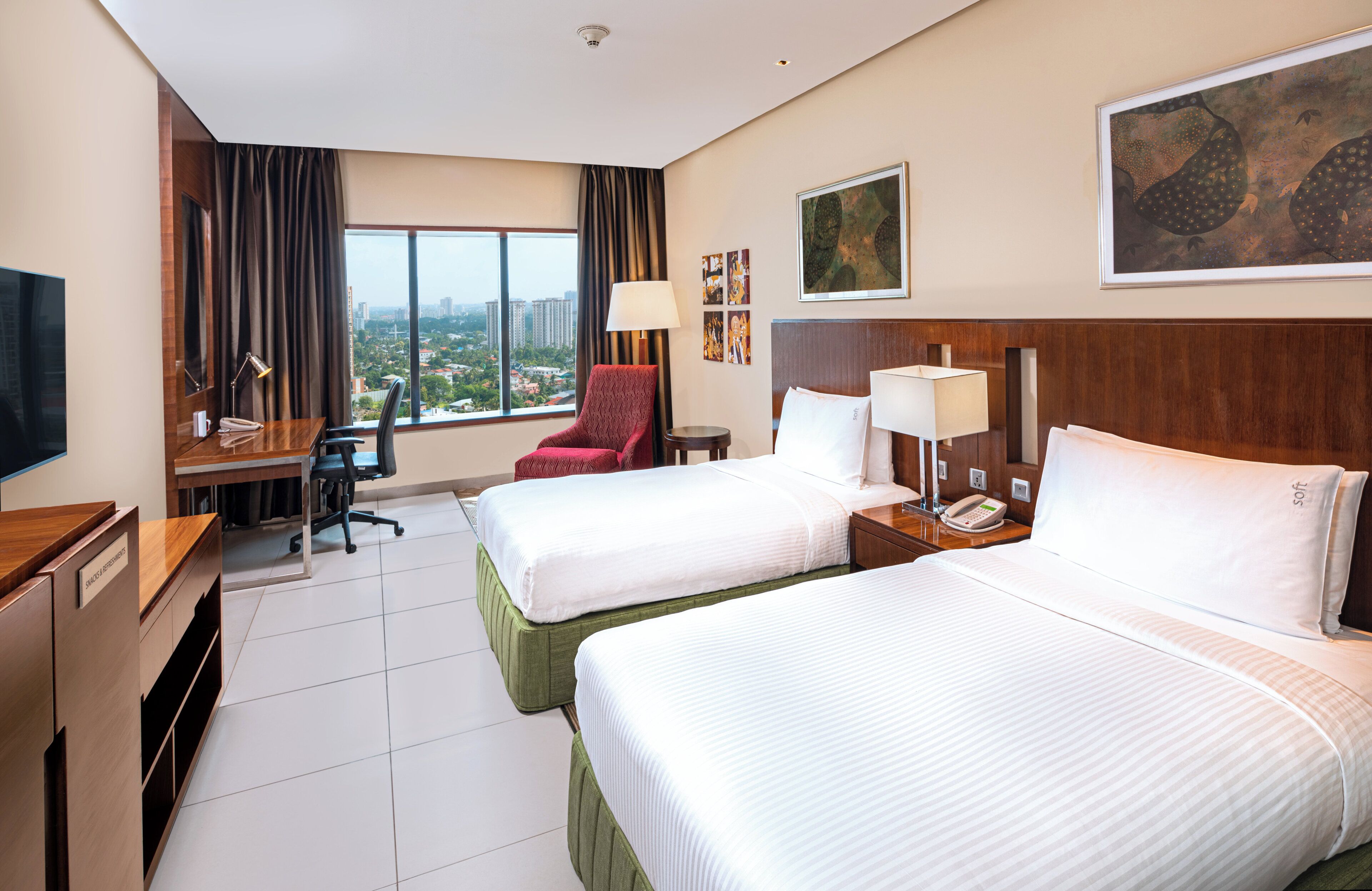 A hotel room with two beds, a desk with a chair, a TV, and a large window with a city view.