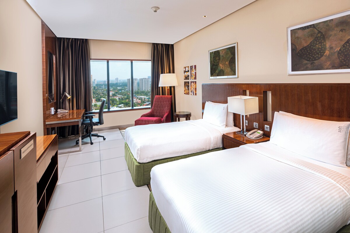 A hotel room with two beds, a desk with a chair, a TV, and a large window with a city view.