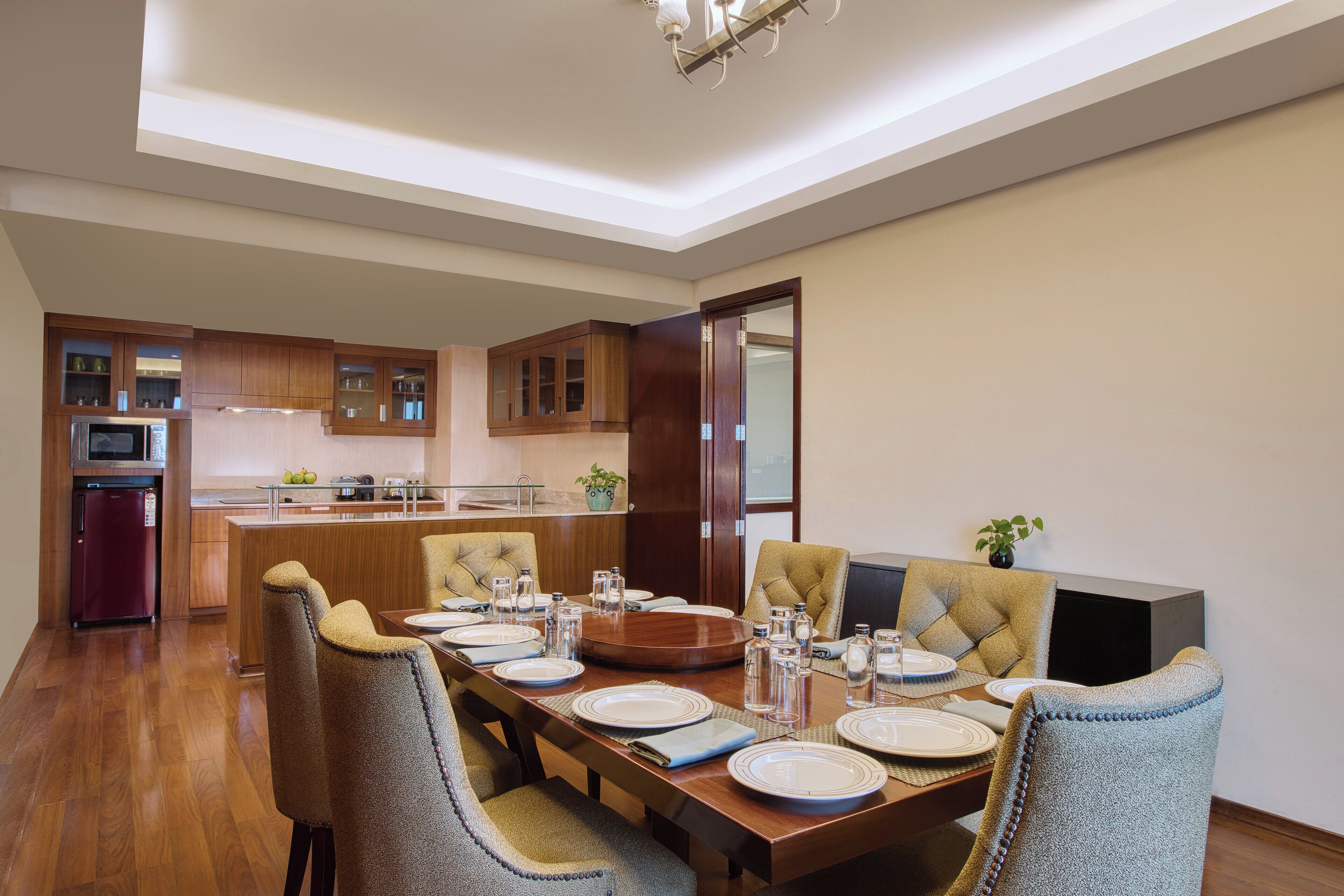 A modern dining area with a wooden table set for six, surrounded by tufted chairs. A kitchen with wooden cabinets and a refrigerator is visible in the background.