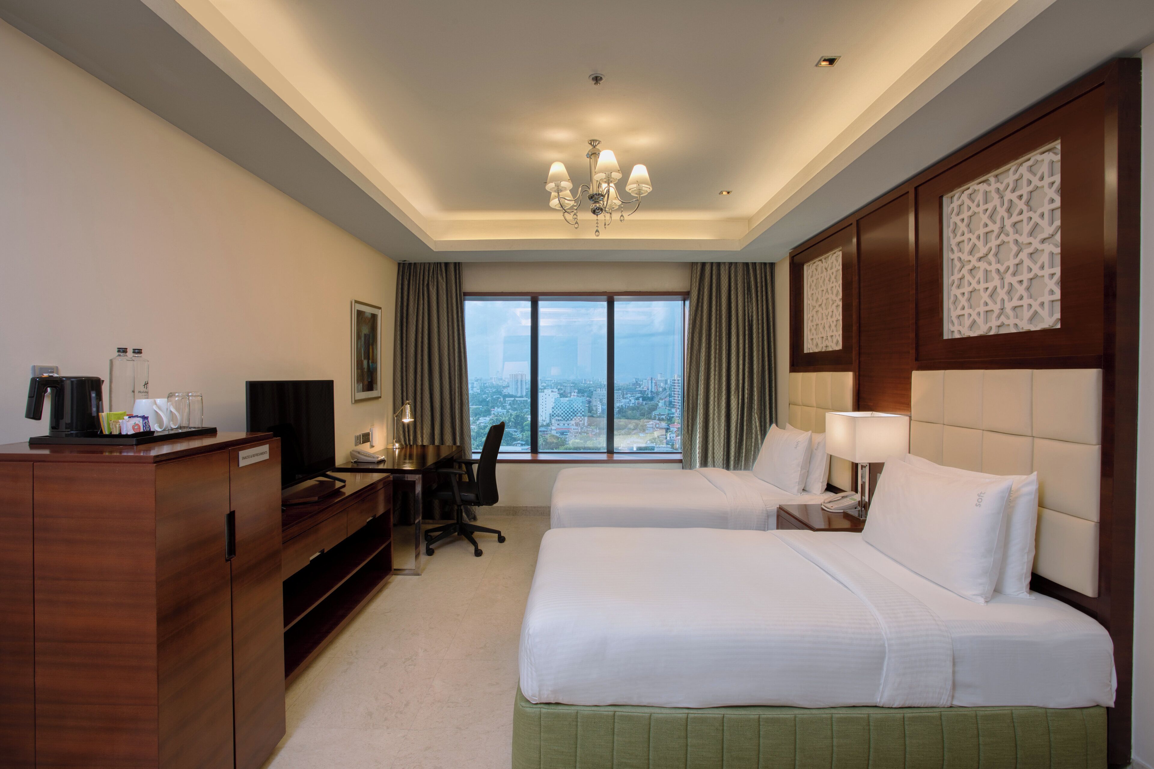 A hotel room with two beds, a desk with a chair, a television, and a view of the city through large windows.