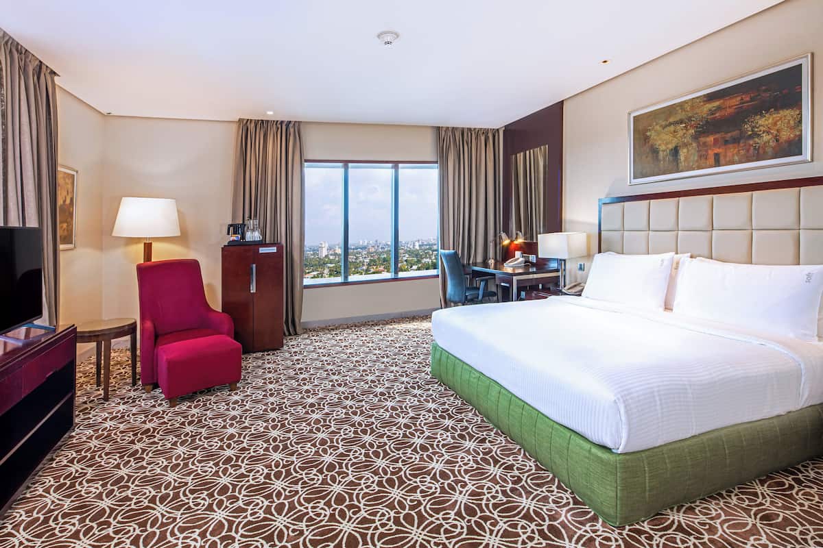 Ein Hotelzimmer mit einem großen Bett, einem Schreibtisch mit Stuhl, einem Fernseher und Blick auf die Stadt durch große Fenster.