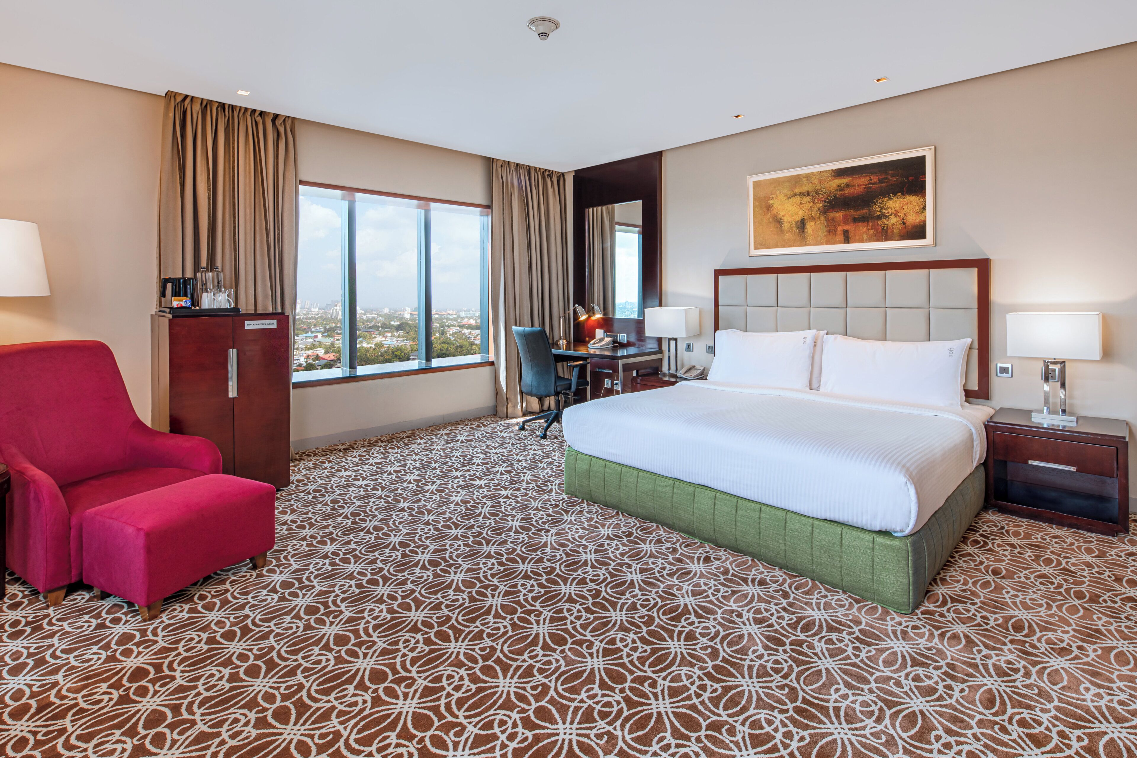 Ein Hotelzimmer mit einem großen Bett, einem Schreibtisch, einem roten Sessel und Blick auf die Stadt durch große Fenster.