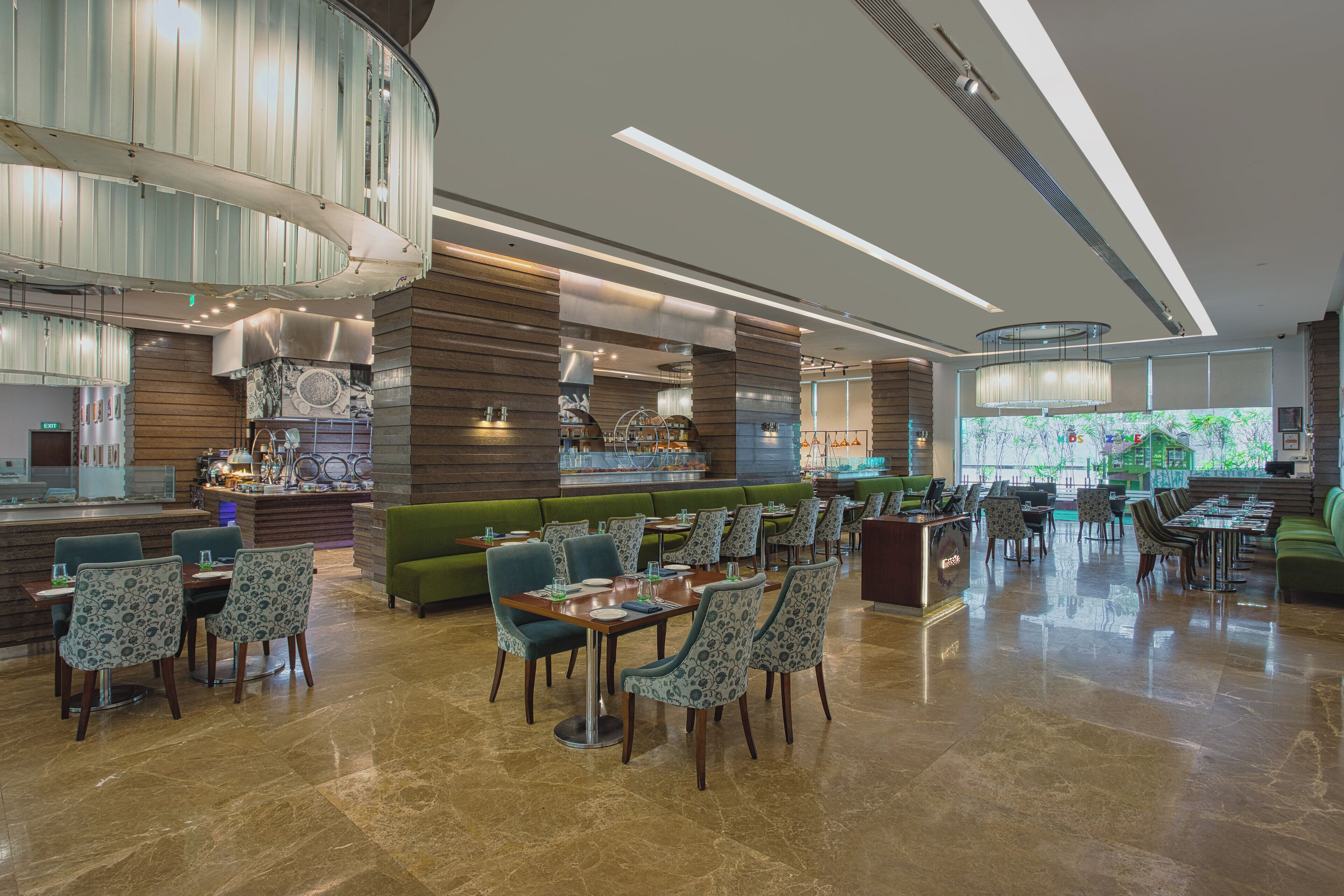 A modern restaurant with a central bar, green seating, and a large window offering a view.