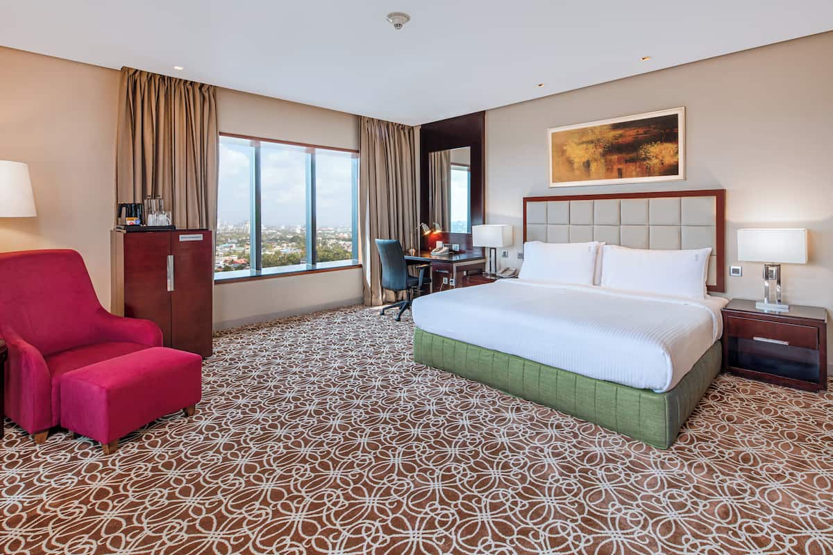 Ein Hotelzimmer mit einem großen Bett, einem Schreibtisch, einem roten Sessel und Blick auf die Stadt durch große Fenster.