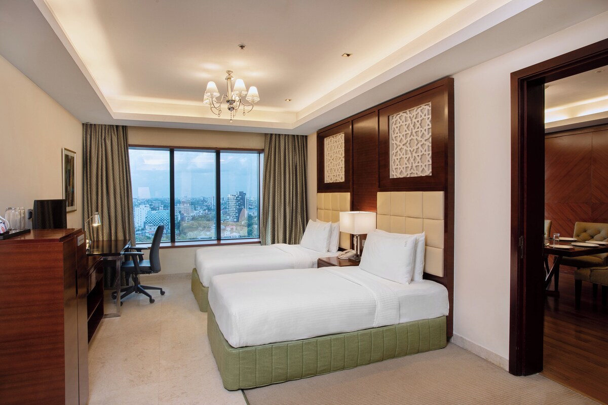 A hotel room with two beds, a desk with a chair, a desk lamp, a telephone, and a view of the city through large windows.