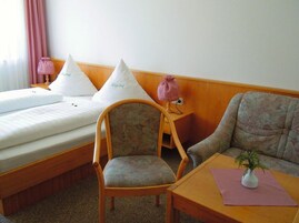 A hotel room with a bed, two pillows, a chair, a sofa, a small table, a lamp, and a plant.