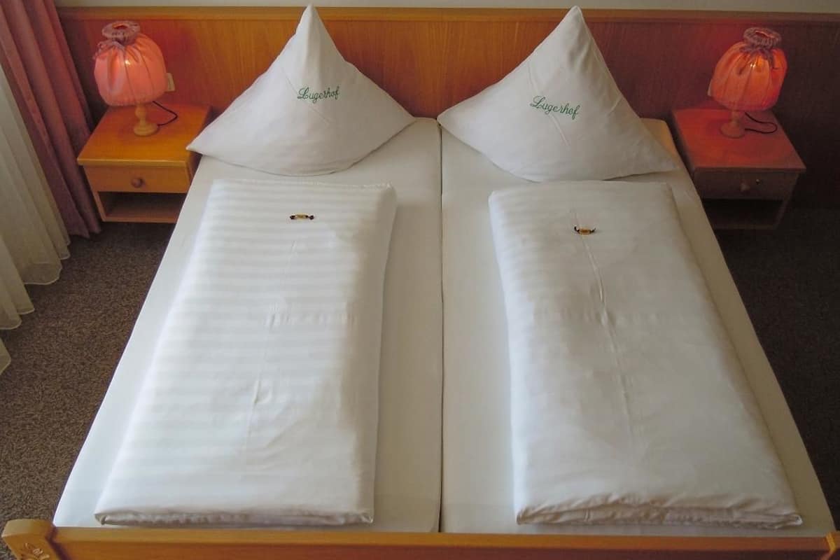 A double bed with two pillows labeled "Lugerhof" and two bedside lamps on wooden nightstands.
