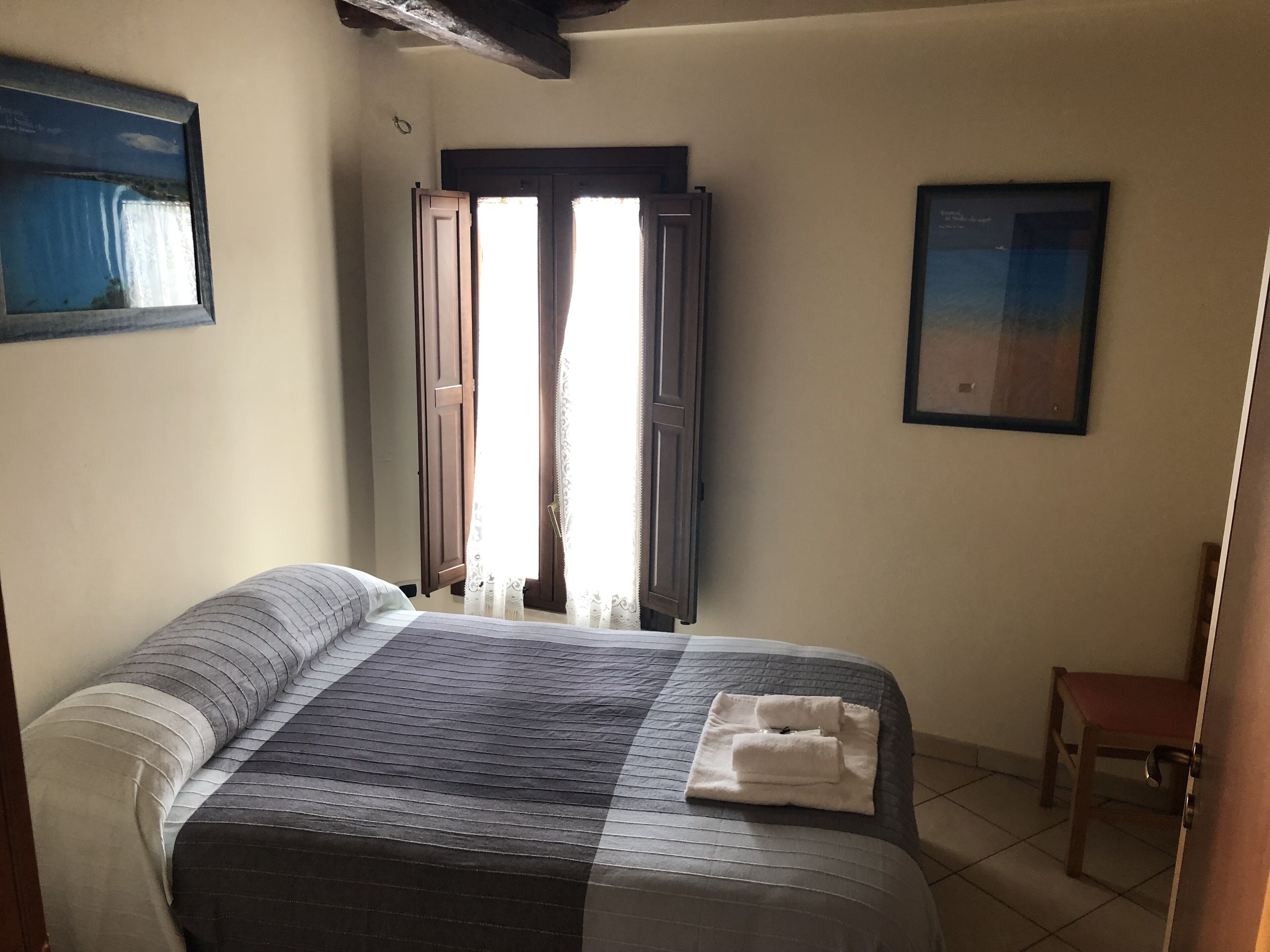 A bedroom with a bed, a wooden chair, a window with curtains, and a framed picture on the wall.