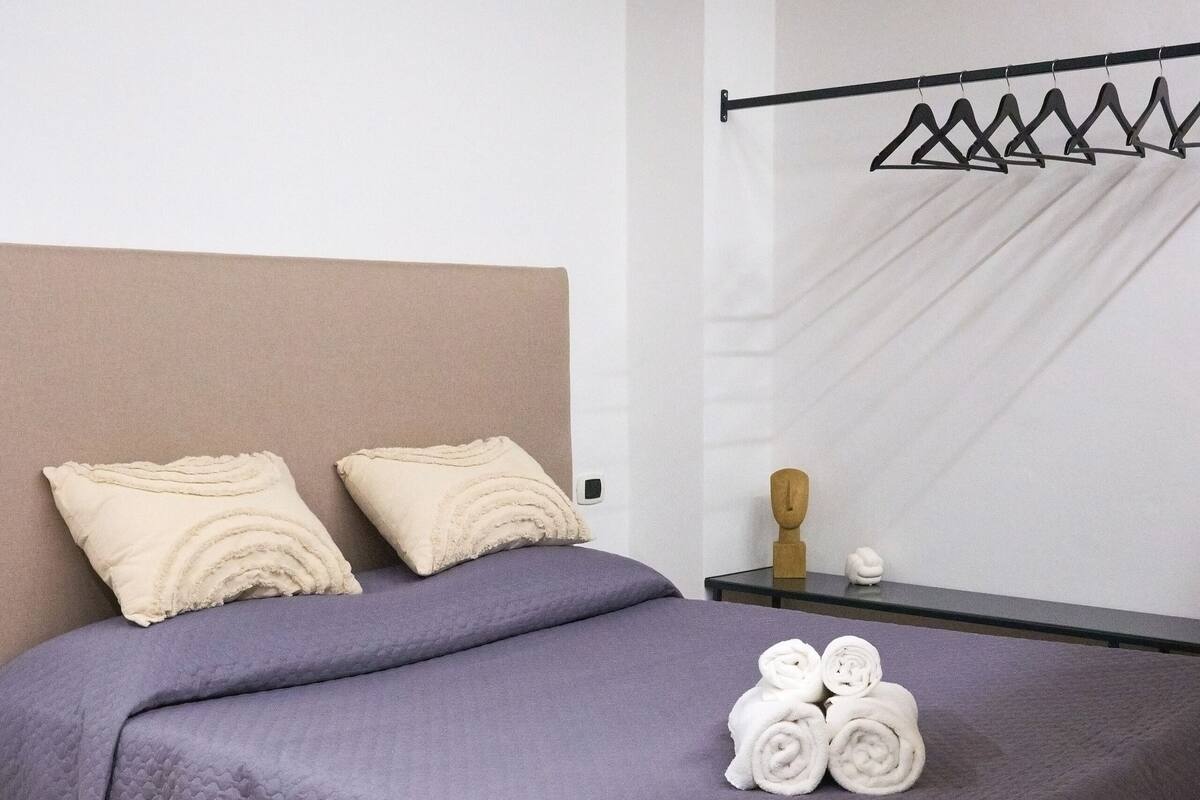 A neatly made bed with a quilted purple blanket, two beige pillows, and a set of rolled towels on the side.