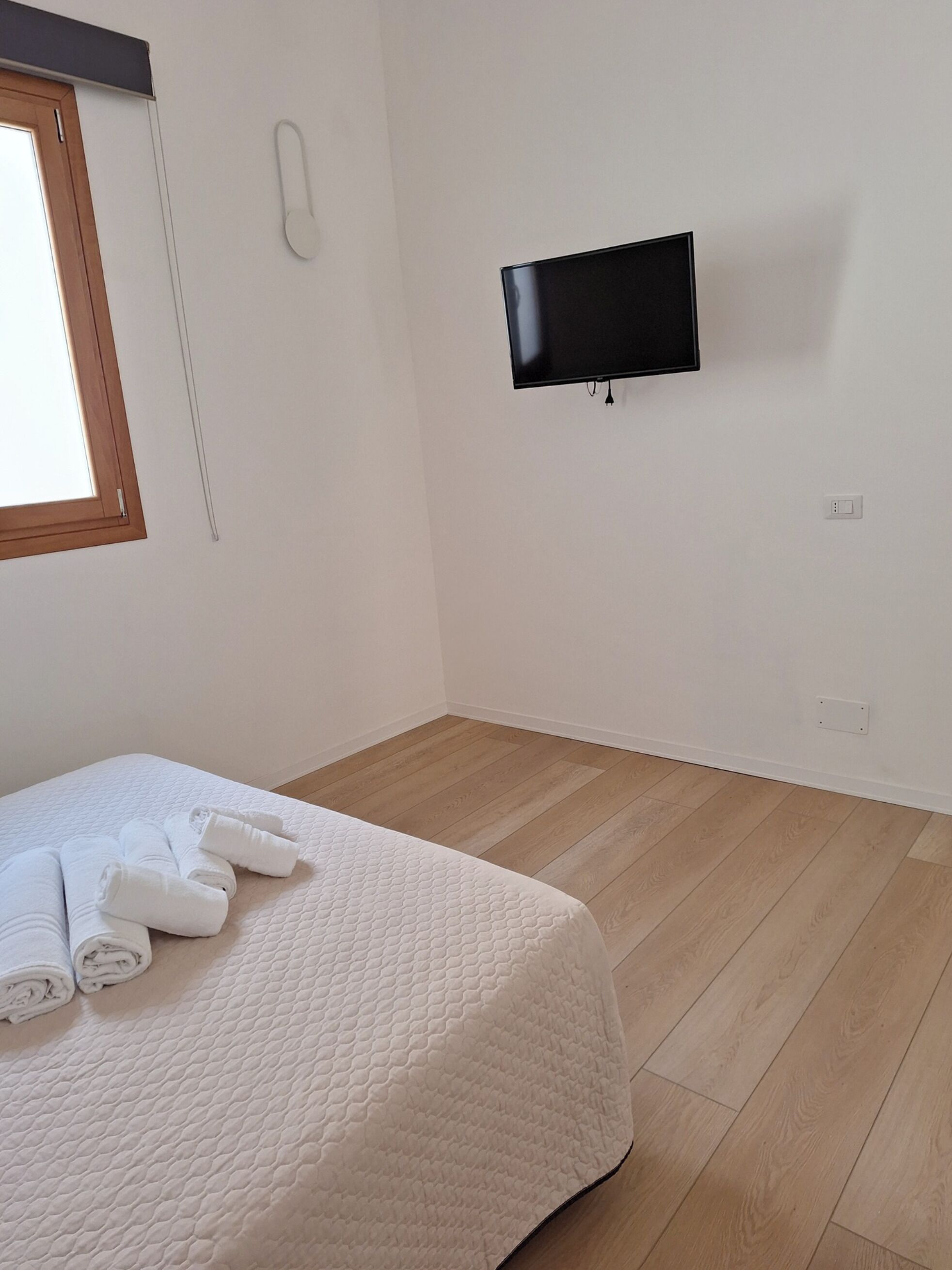 A bedroom with a bed, a flat-screen TV mounted on the wall, a telephone, and a window with blinds.