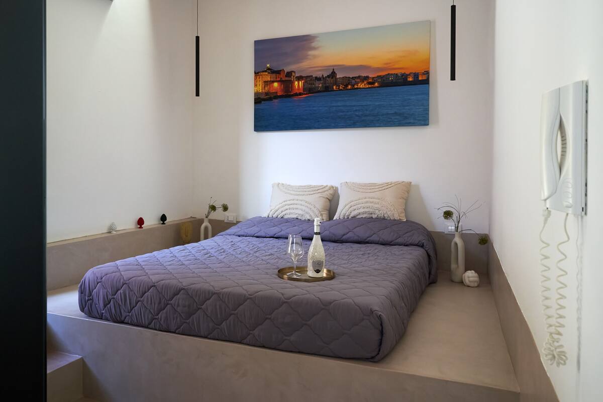 A modern bedroom with a large bed, a cityscape painting, and a bedside table with a vase.