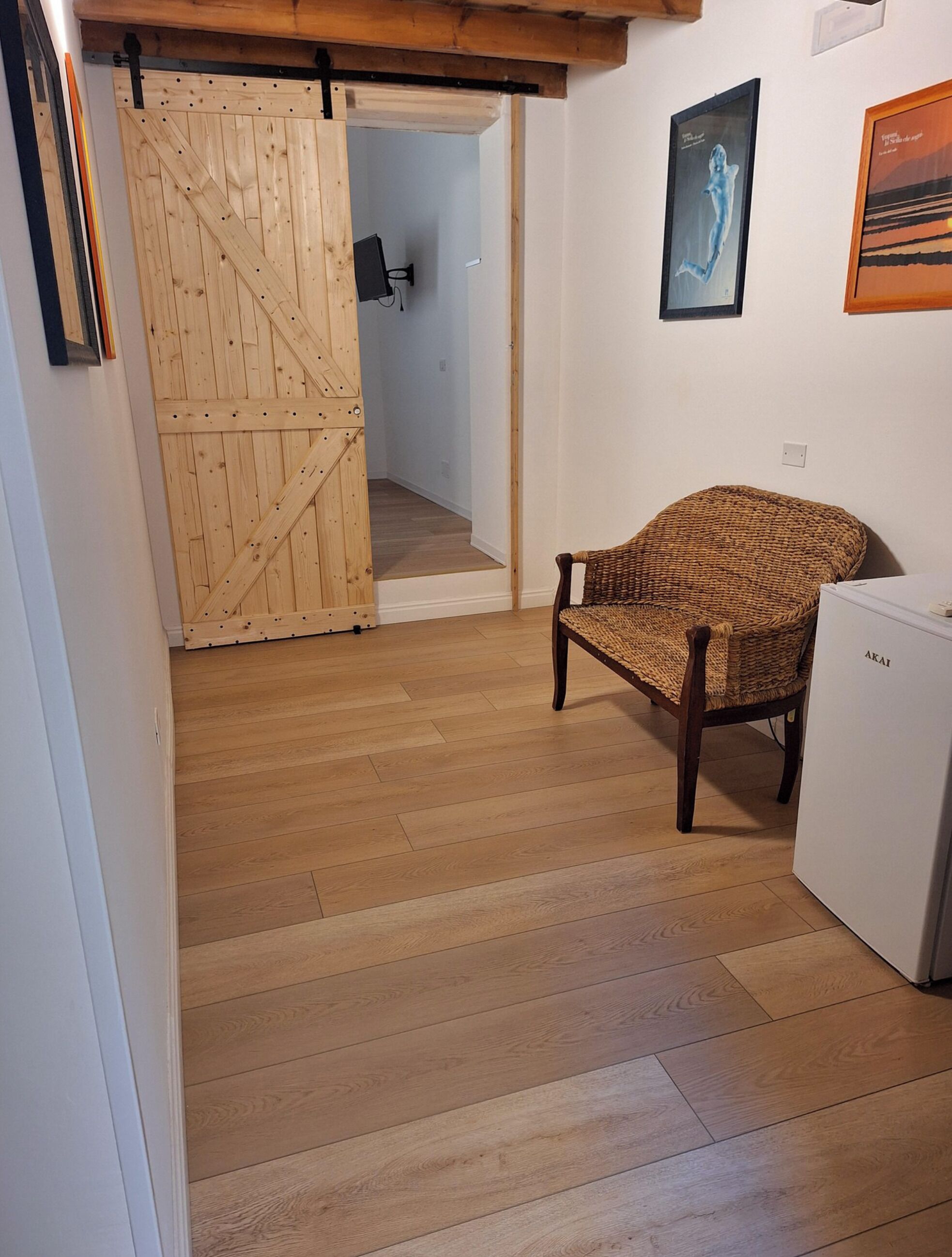 A room with a wooden floor, a wicker chair, a white refrigerator, and a wooden door.