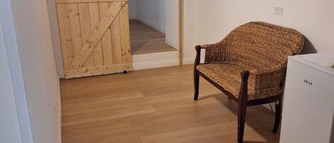 A room with a wooden floor, a wicker chair, a white refrigerator, and a wooden door.