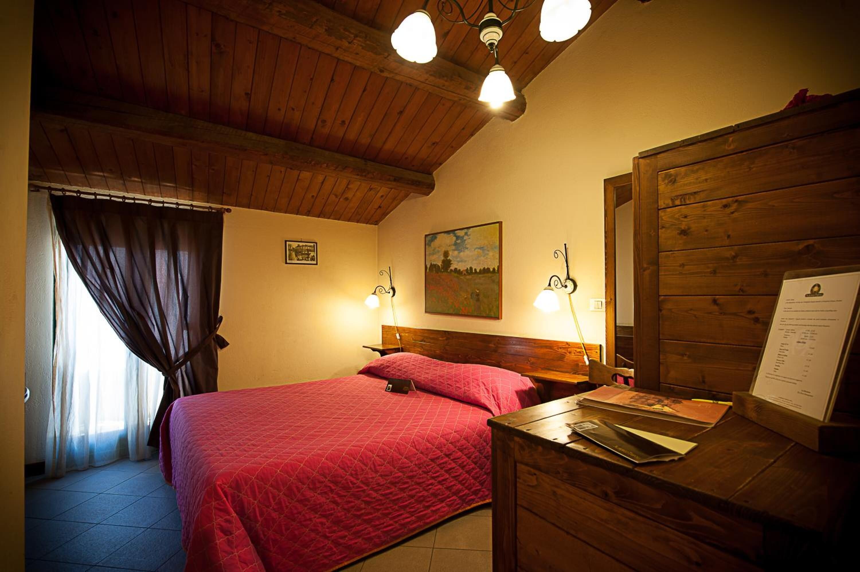 A bedroom with a wooden ceiling, a bed with a red quilt, two bedside lamps, a wooden nightstand, and a wall-mounted painting.