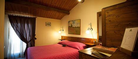 A bedroom with a wooden ceiling, a bed with a red quilt, two bedside lamps, a wooden nightstand, and a wall-mounted painting.
