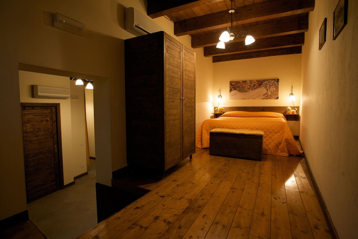 Un dormitorio con suelo de madera, una cama con colcha naranja, una mesita de noche y un cuadro en la pared.