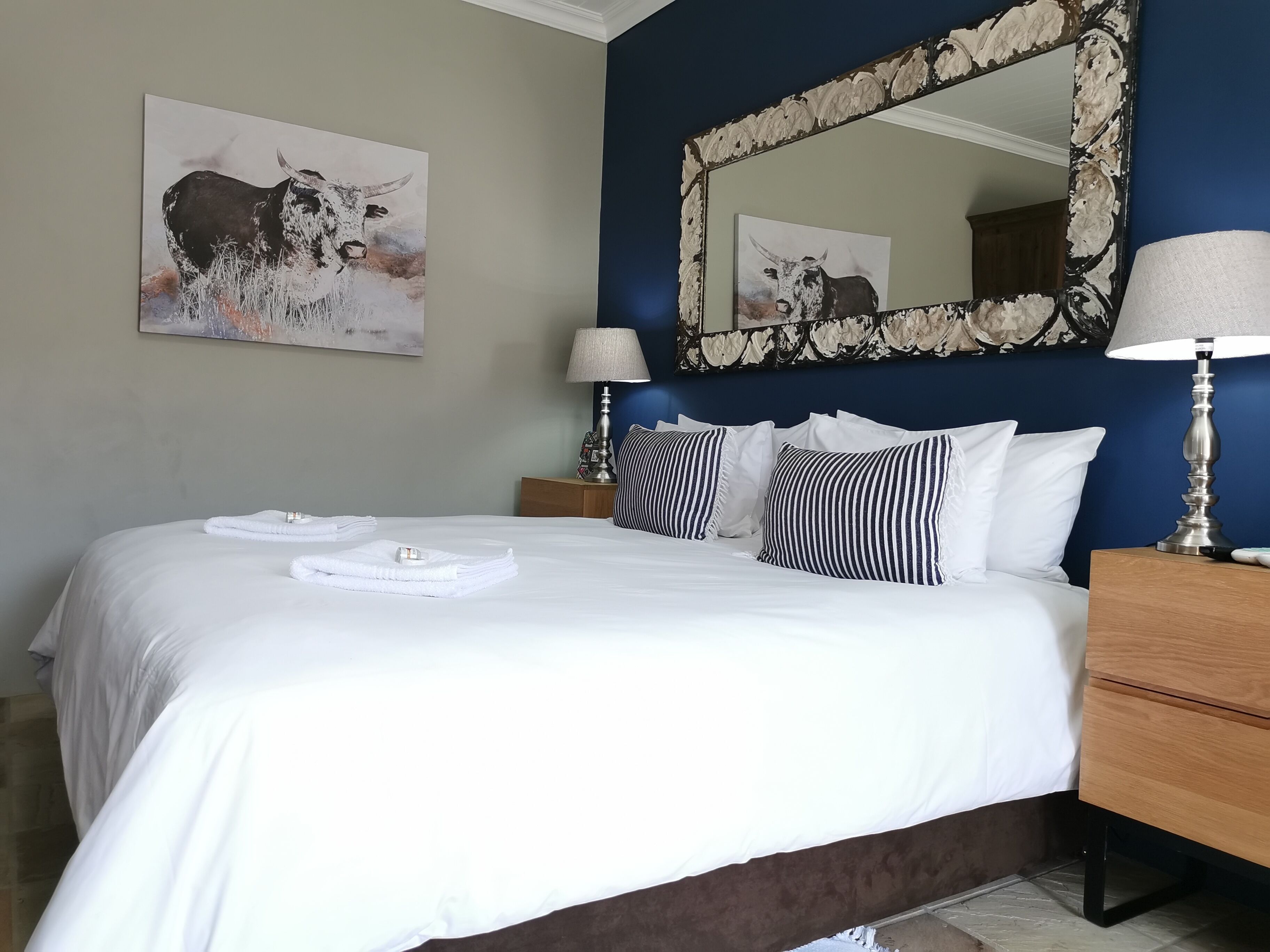 A neatly made bed with white linens and a folded towel, a bedside table with a lamp, and a mirror with a decorative frame.