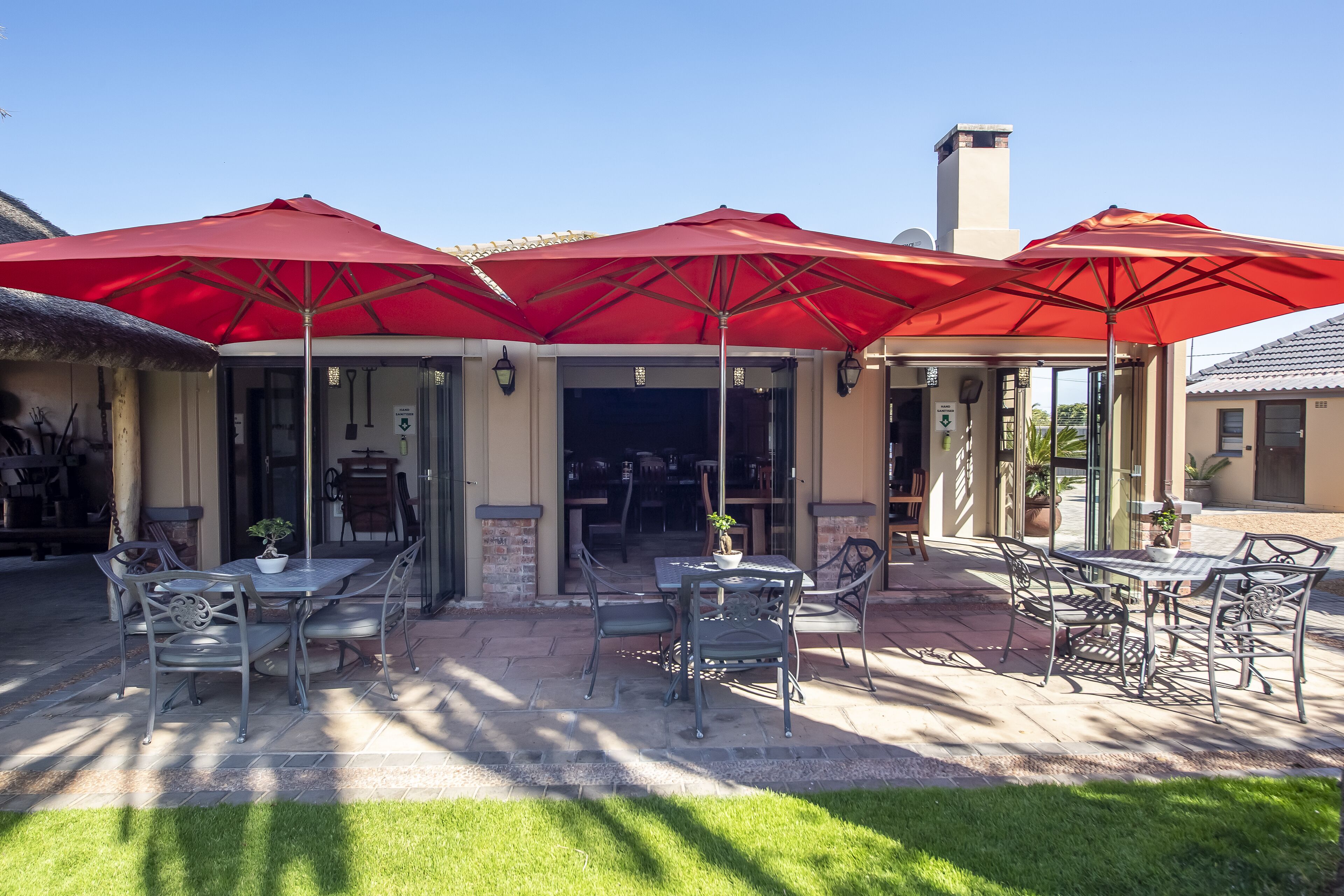 An outdoor patio area with tables and chairs under red umbrellas, a brick wall, and a glass door entrance.
