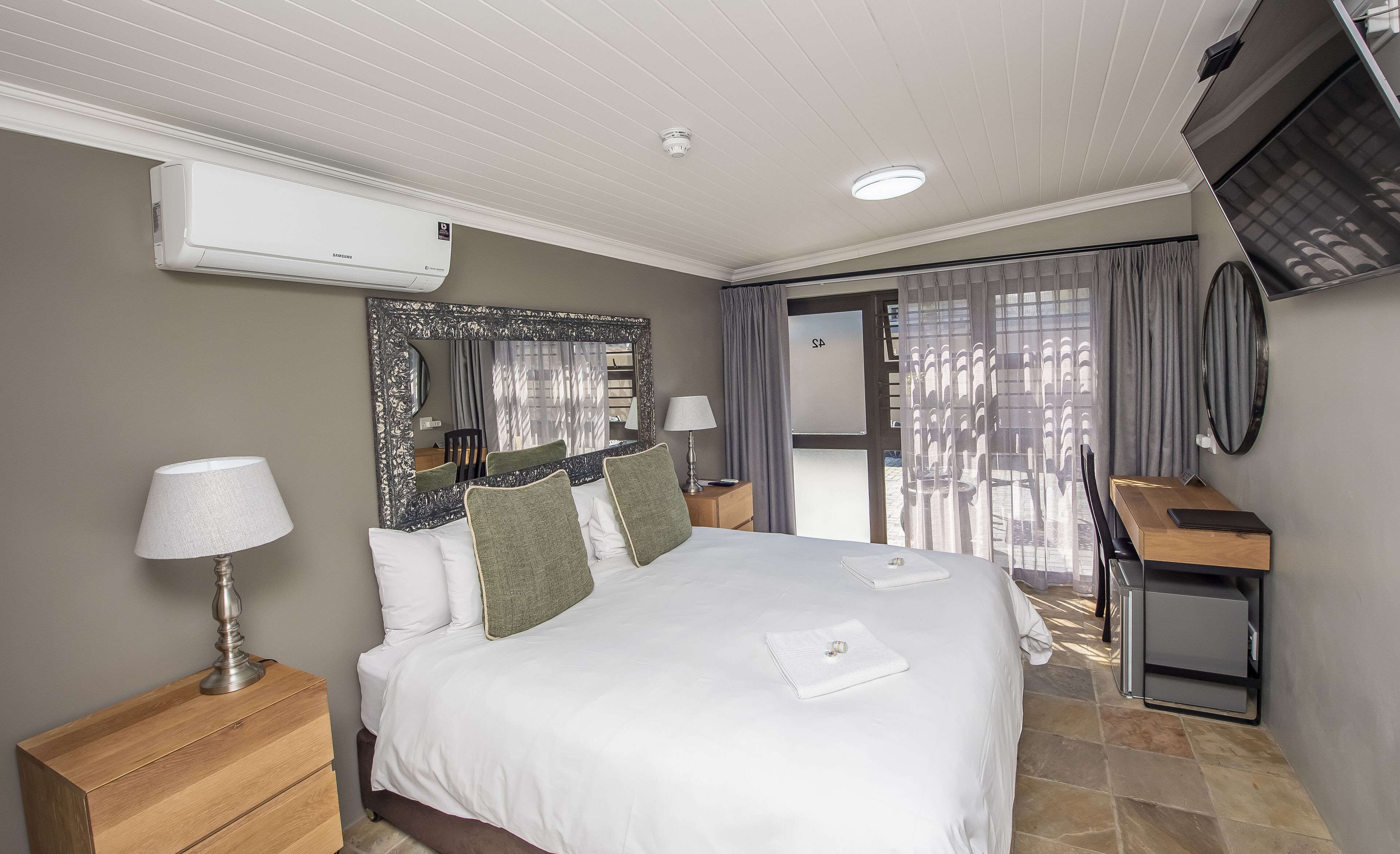 A hotel room with a bed, bedside tables, a mirror, a desk, and an air conditioning unit.