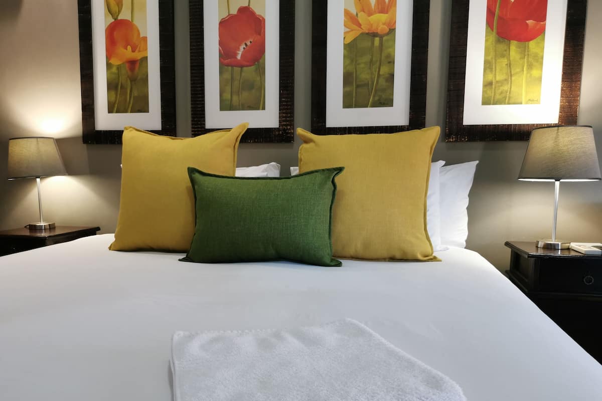 Cama bem arrumada com toalha branca, três almofadas em amarelo e verde, e uma obra de arte na parede.