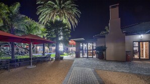 A well-lit outdoor area with a paved walkway, palm trees, and outdoor seating under red umbrellas. - Excellent Guest House (Cape Town)