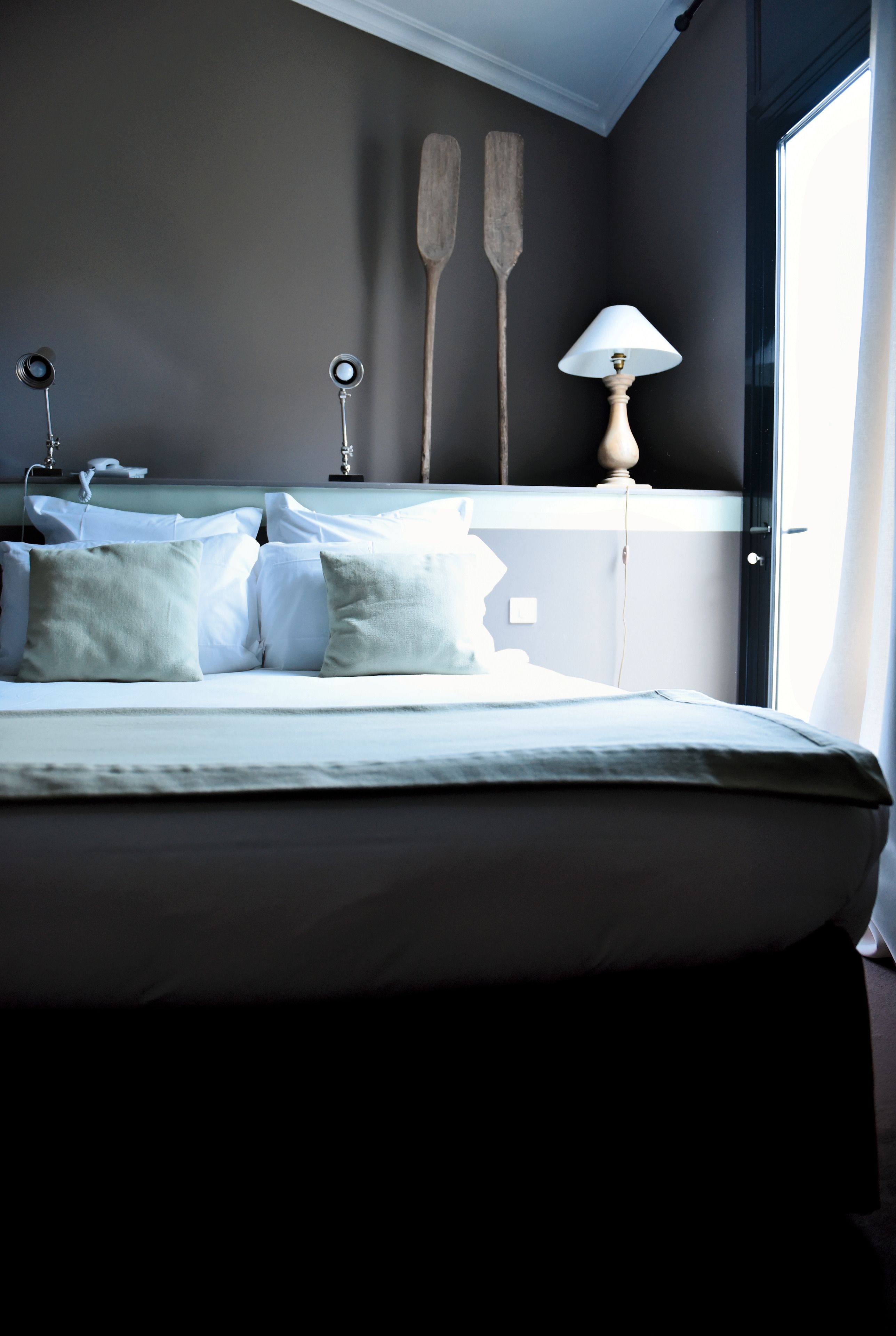 A neatly made bed with white linens and pillows, a bedside lamp, and wooden paddles on a wall shelf.