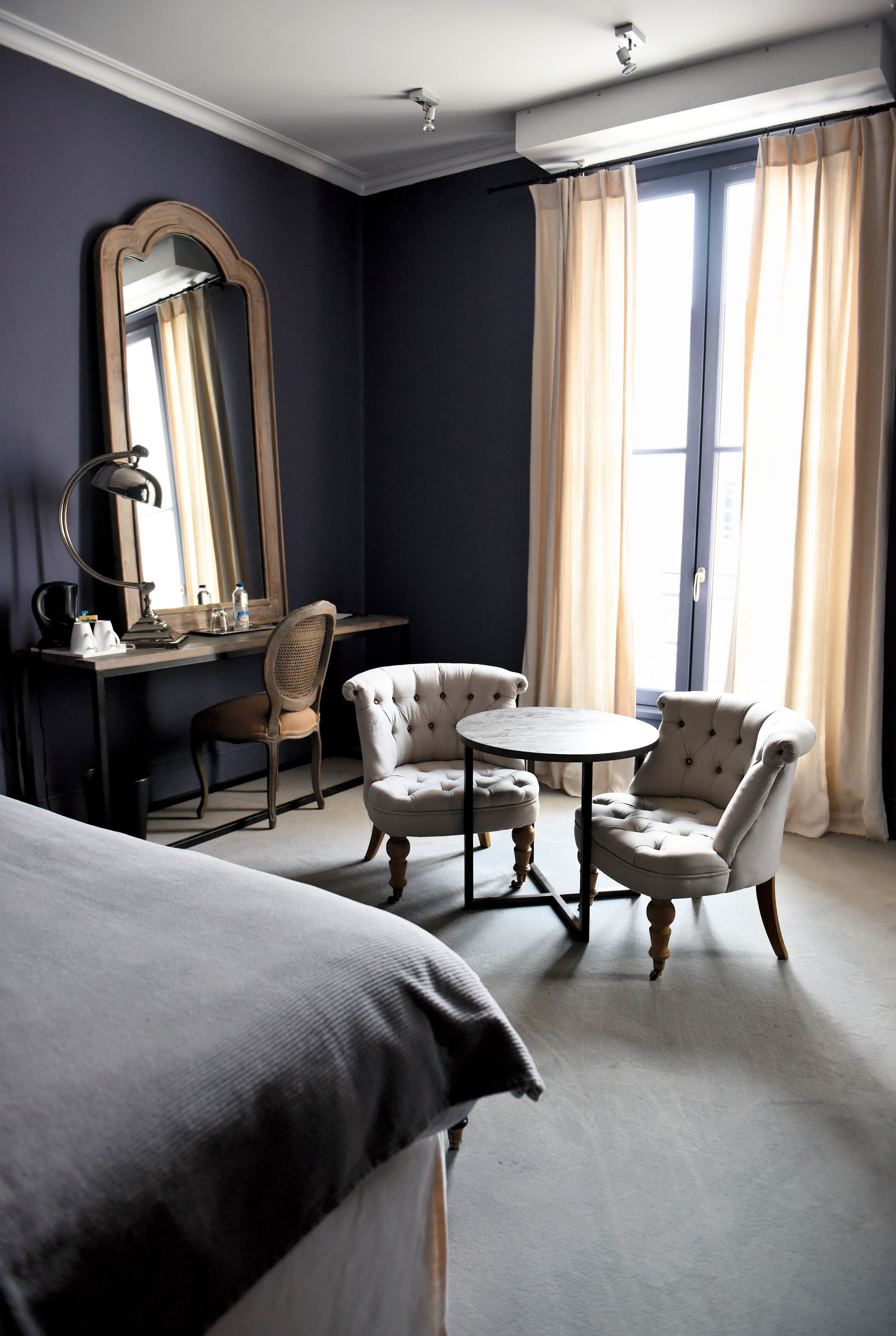 Une chambre d’hôtel comprenant un lit, un bureau avec une chaise, une table ronde avec deux chaises, un grand miroir et une fenêtre avec des rideaux.