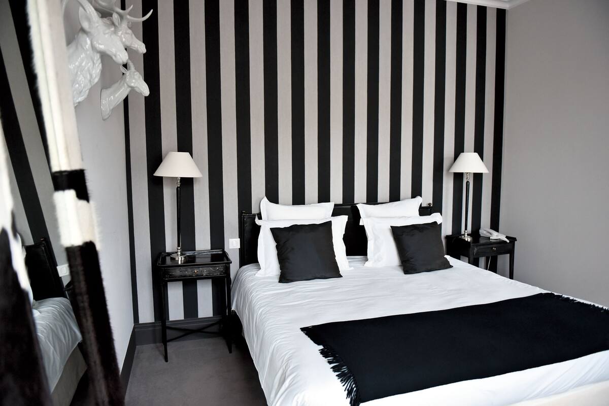 Una habitación de hotel moderna con una cama grande, pared a rayas blancas y negras, dos mesitas de noche con lámparas y una cabeza de venado decorativa en la pared.