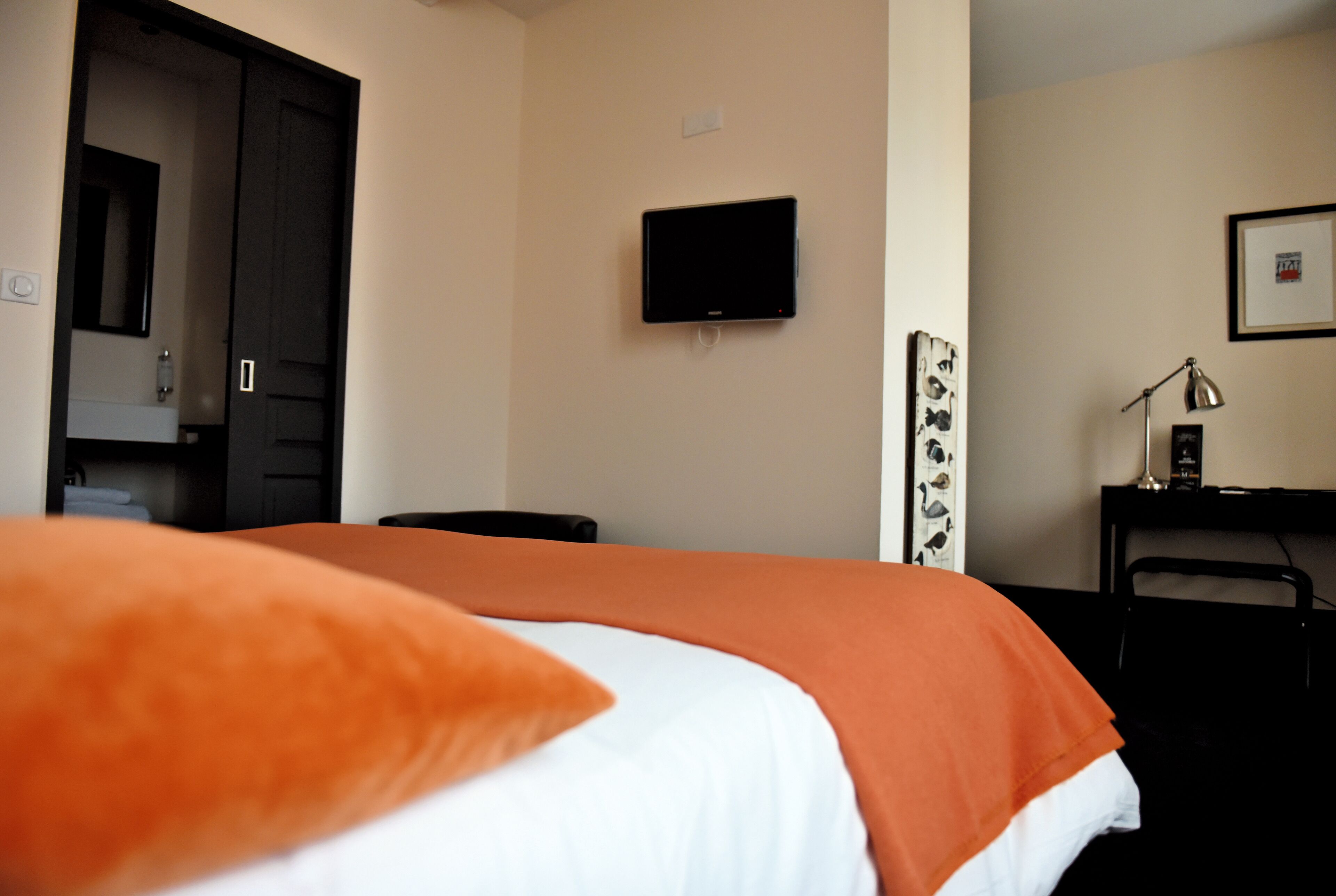 A hotel room with a bed, a television mounted on the wall, a desk with a lamp, and a chair.