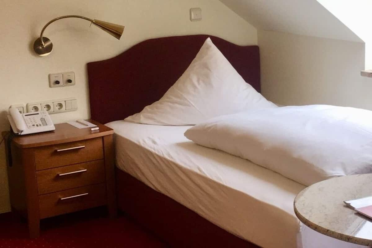 A single bed with white bedding, a bedside table with a lamp, a small round table with a magazine, and a skylight above the bed.