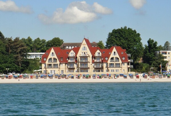 A large hotel with multiple red-roofed buildings, a beach in the foreground, and a body of water in the background.
