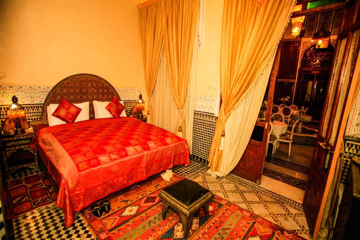 Une pièce traditionnelle marocaine avec un sol à carreaux, un lit recouvert d’une courtepointe rouge et un petit tabouret en bois.