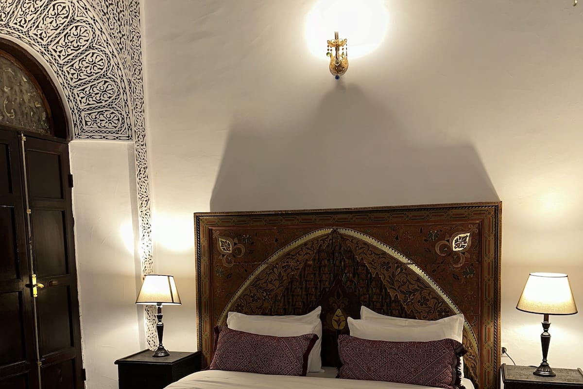 Un lit bien fait, avec une literie blanche et des coussins à motifs, flanqué de deux tables de chevet avec des lampes, dans une pièce aux décorations murales élaborées et dotée d’une porte en bois.