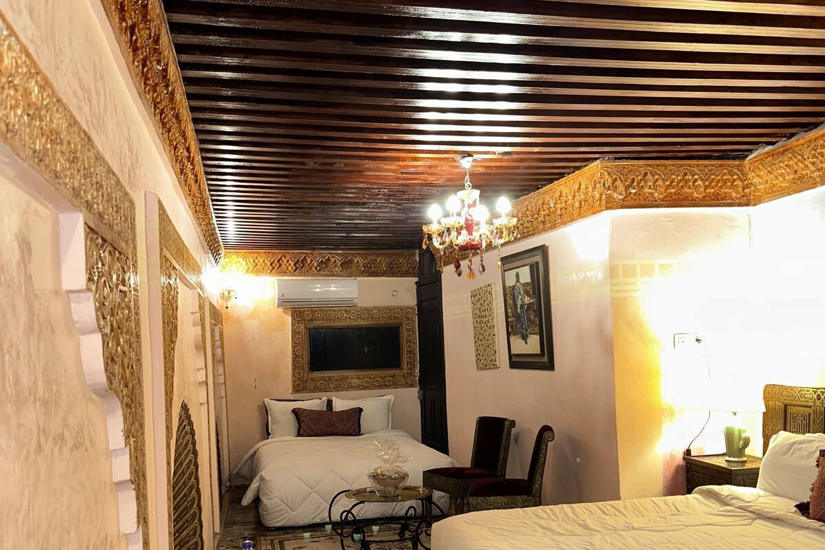 Une chambre de style traditionnel, avec un grand lit, une petite table sur laquelle se trouvent une bouteille et des verres, un fauteuil décoratif et un lustre.