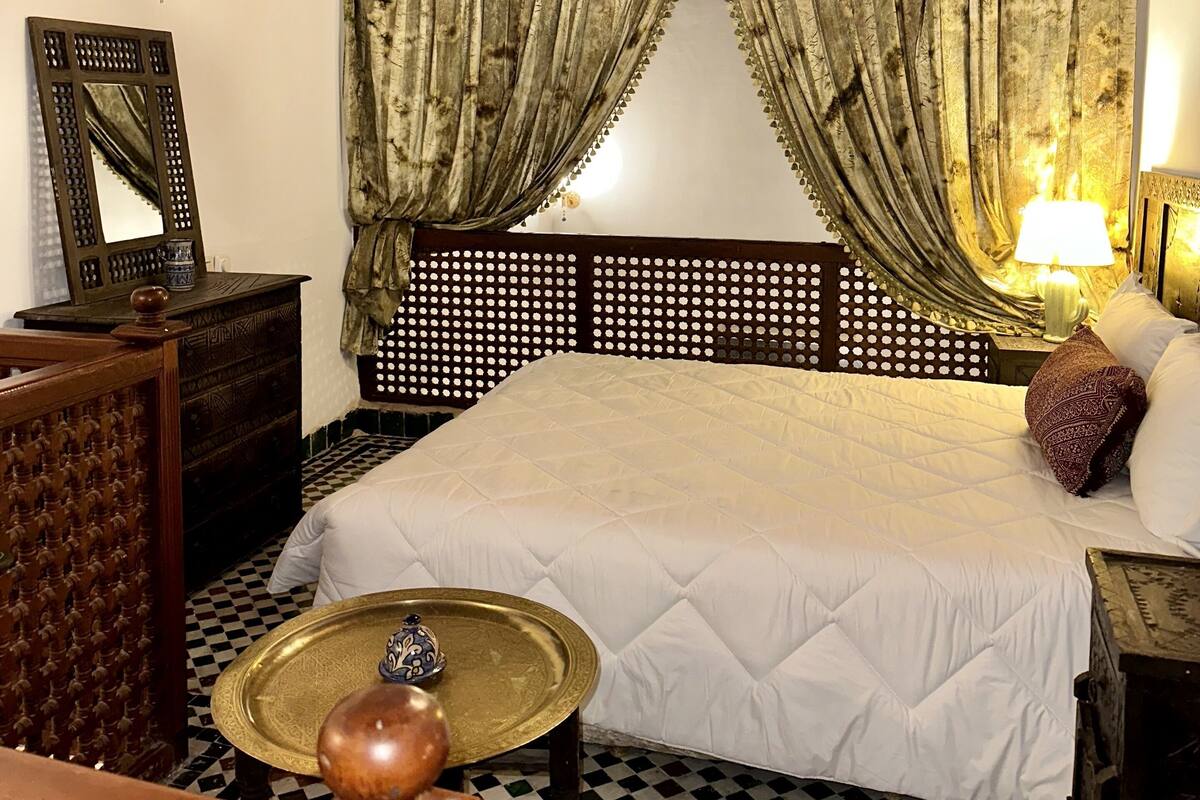 Une chambre à coucher comprenant un lit, une tête de lit en bois, des rideaux, un lustre, une lampe, un miroir et un plateau décoratif.