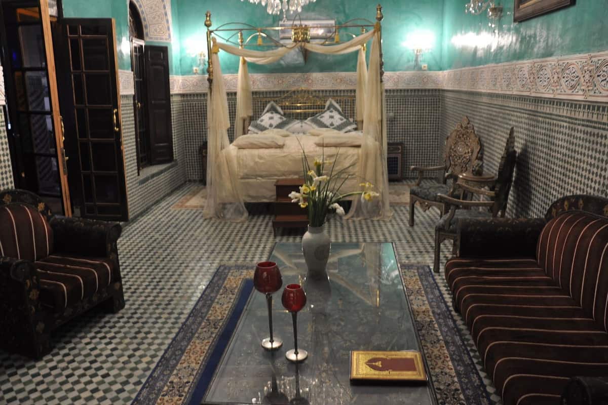 Une chambre avec un lit à baldaquin, un lustre, un vase rempli de fleurs et deux verres à vin rouge posés sur une table.