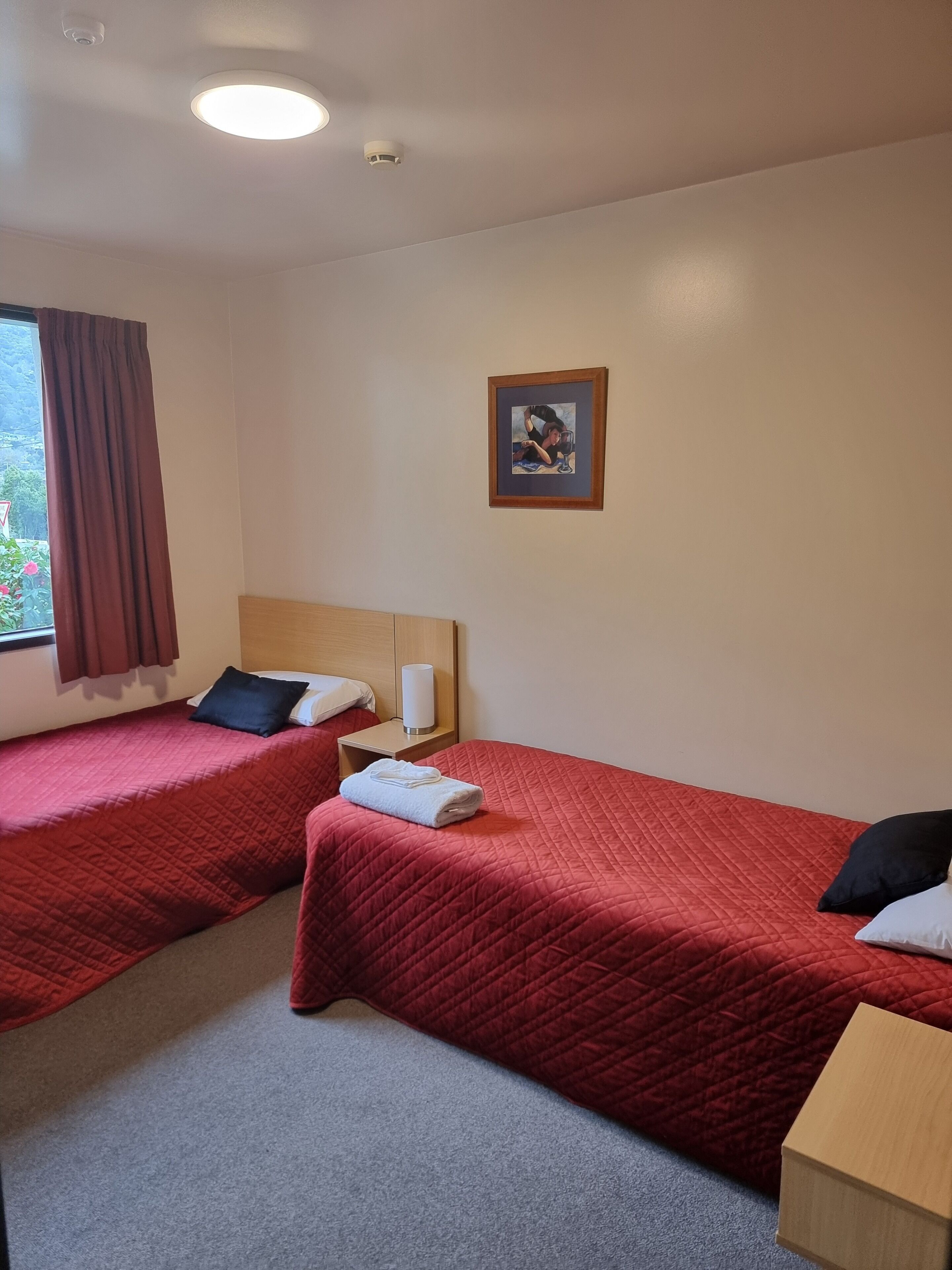 A hotel room with two single beds, each with a red quilted bedspread and pillows, a wooden headboard, a small bedside table with a lamp, and a framed picture on the wall.