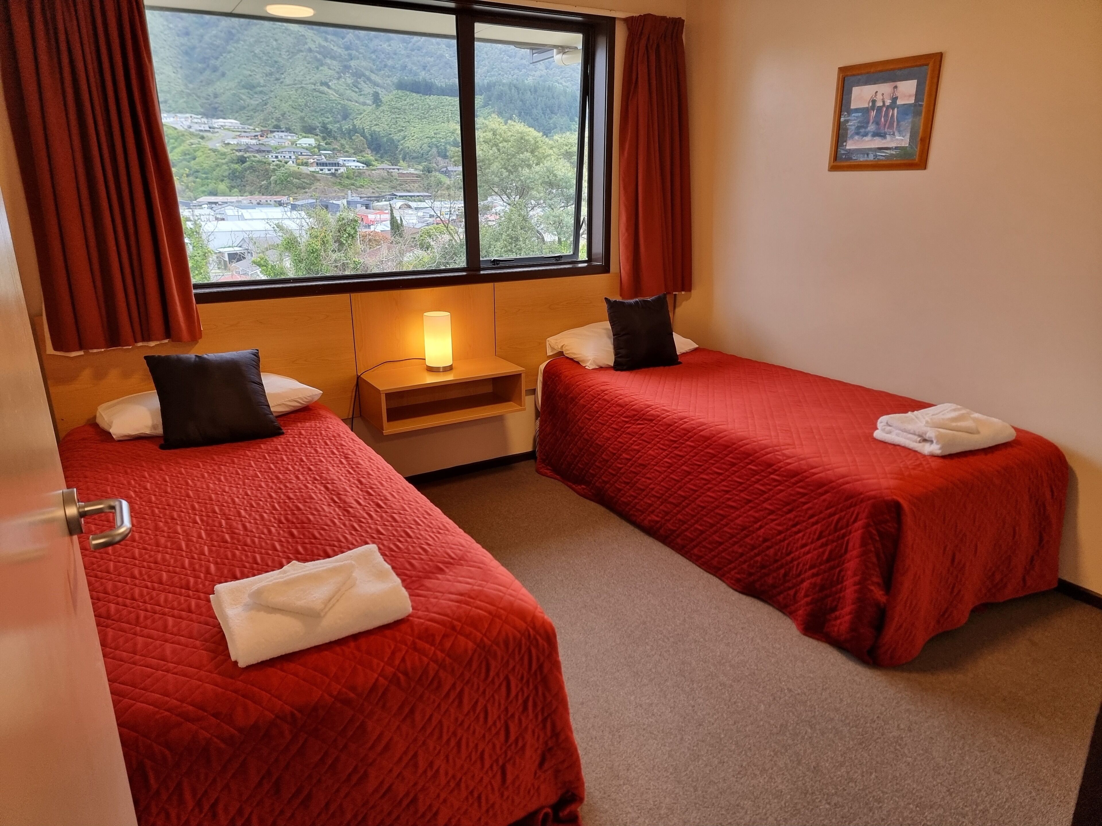 Habitación de hotel con dos camas, una mesita de noche, una lámpara, una ventana con cortinas y un cuadro en la pared.