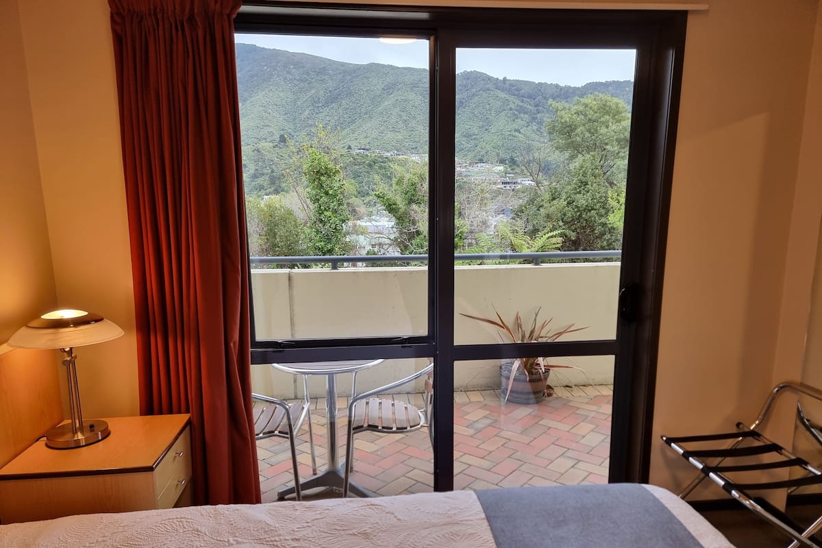 A hotel room with a bed, a desk, a lamp, a potted plant, and a view of a balcony with a table and chairs.